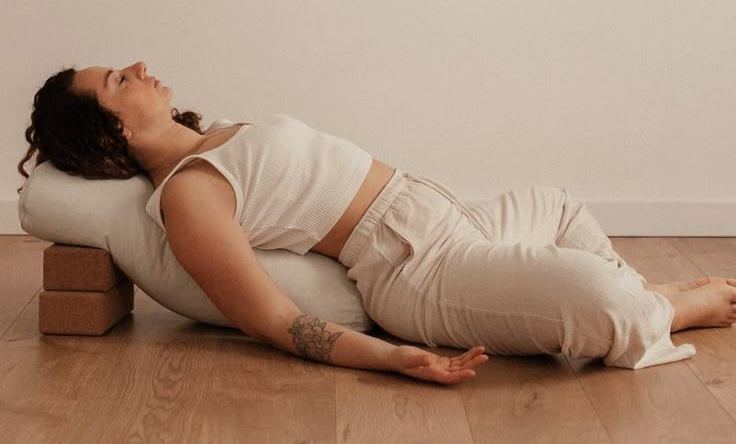 Woman in cream athletic wear in peaceful restorative yoga pose with props on wooden floor